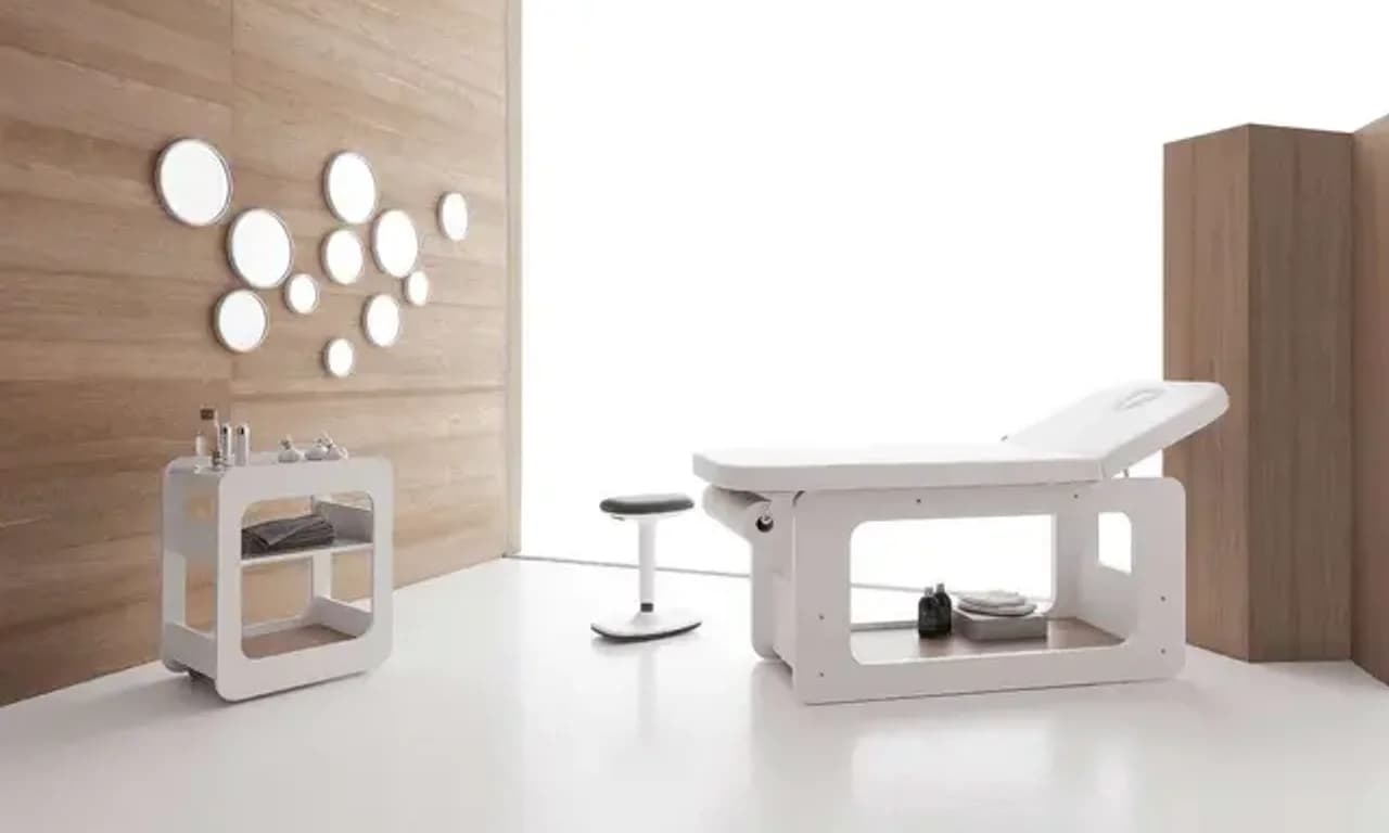 Cabina estetica arredata: lettino bianco con vano aperto, carrello di servizio e specchi rotondi su parete in legno