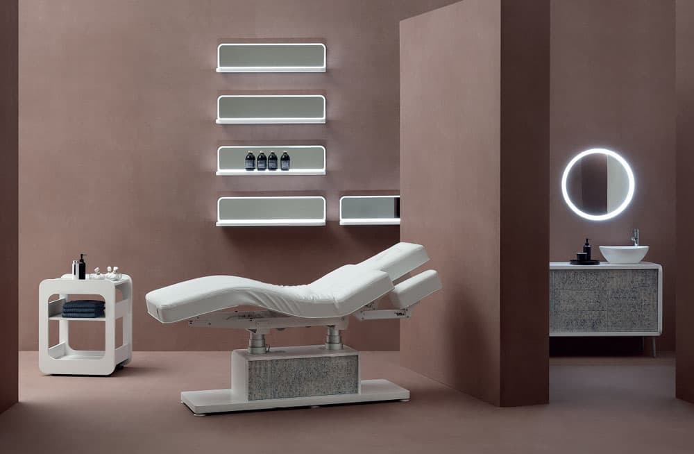 Cabina estetica arredata Vismara: lettino bianco, carrello portaoggetti, mensole luminose e specchio rotondo su parete color terra